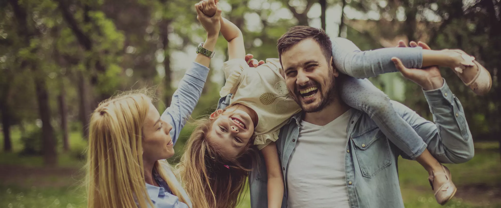A mother, father and young girl are enjoying an English woodland. The father is carrying the young girl on his shoulders..
