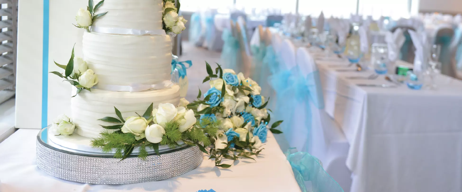 In the foreground a wedding cake is decorated with white and green flowers. In the background a room is set up for a wedding breakfast..