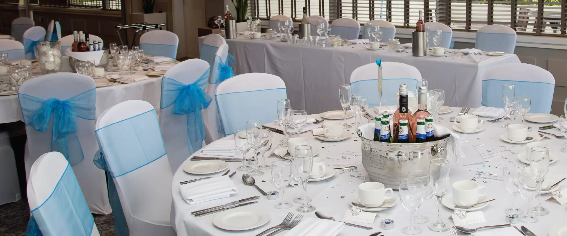 Tables set up for a wedding breakfast with white table and chair covers, blue ribbons and buckets of drinks on the tables..