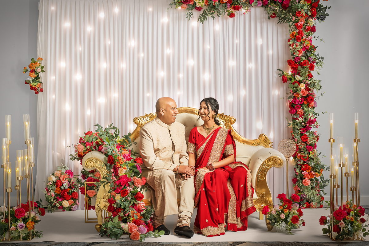 A couple sitting on a white and gold couch. There are red, orange and pink flowers draped over the sofa, draped over a lit backdrop, and tucked into large candle centrepieces to either side..