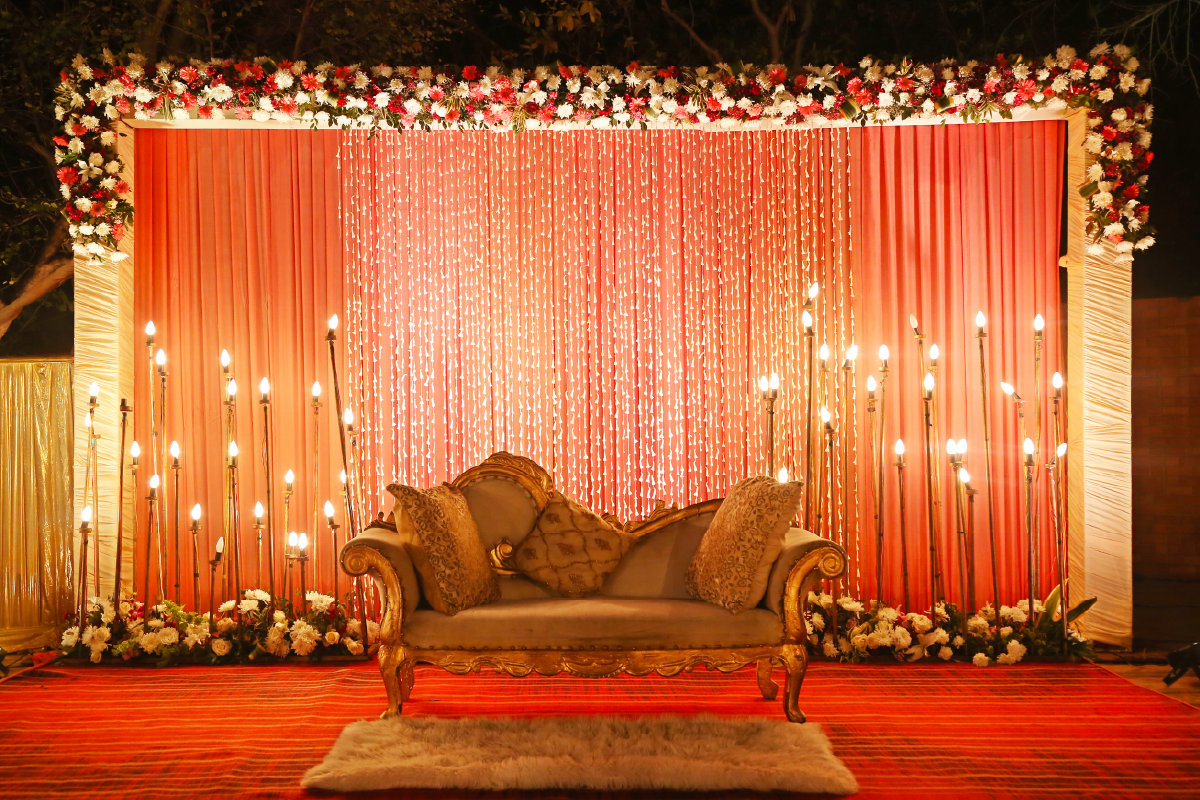 A cream and gold wedding chair set up on a central platform with a flower arch and dozens of lights behind. The backdrop is a bright red/orange.